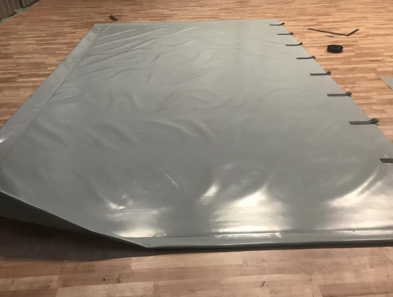 Szara plandeka z uchwytami rozłożona na drewnianej podłodze, obok leżą narzędzia i taśma izolacyjna.