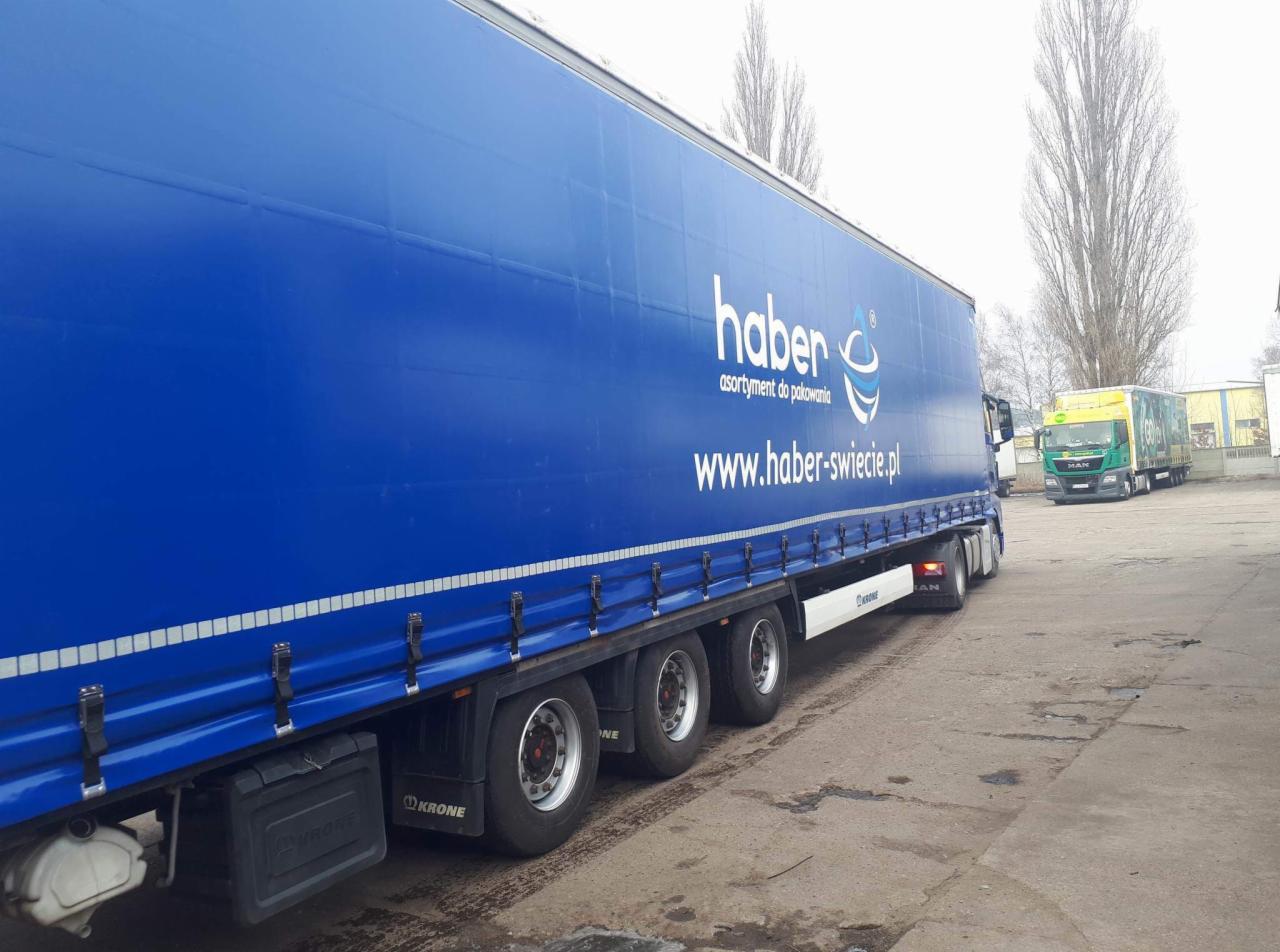 Niebieska naczepa ciężarówki z logo 'haber' i adresem strony internetowej, zaparkowana na placu, w tle inny pojazd ciężarowy.