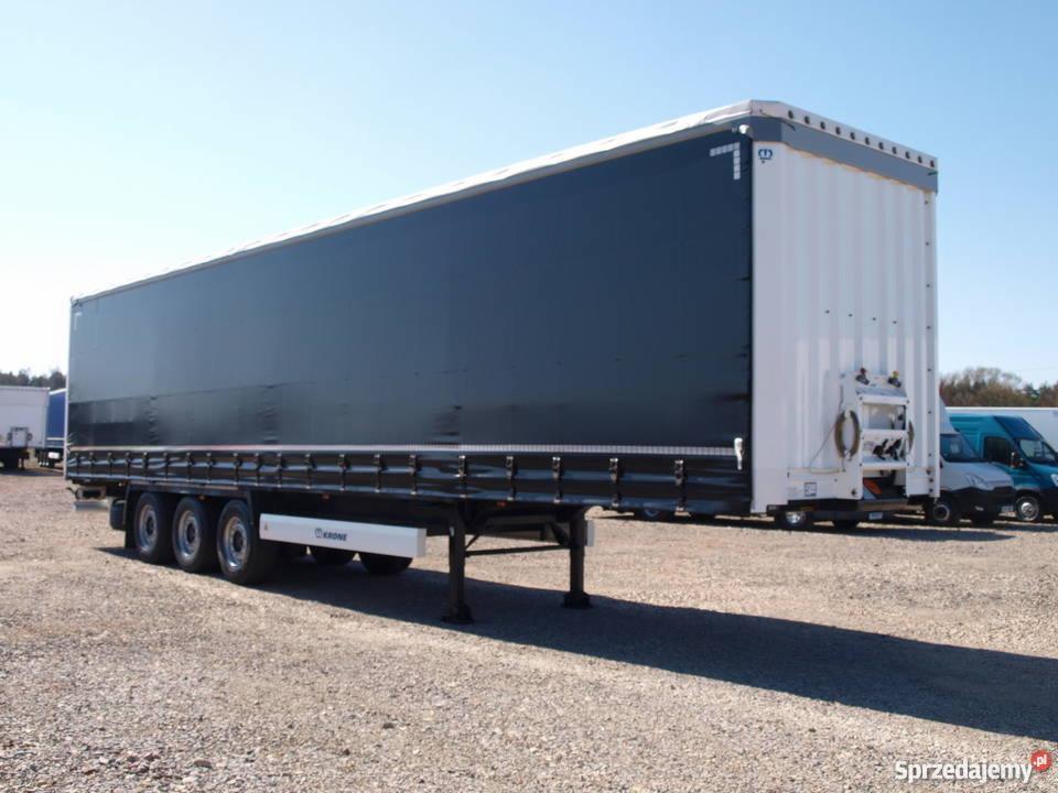 Naczepa ciężarowa marki Krone z czarną plandeką i białym tyłem, zaparkowana na żwirowym placu w słoneczny dzień.