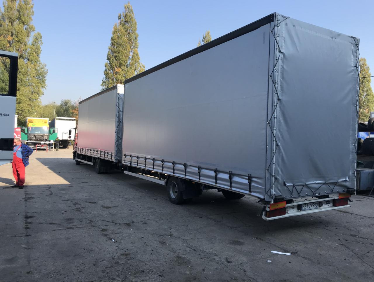 Długi zestaw ciężarowy z szarą plandeką, widoczny pracownik w czerwonym kombinezonie na tle betonowego placu i drzew.