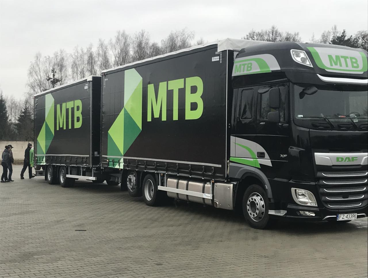 Czarny ciągnik siodłowy DAF XF z naczepą, z dużym logo MTB na burcie, zaparkowany na brukowanym placu, z widocznymi postaciami w oddali.