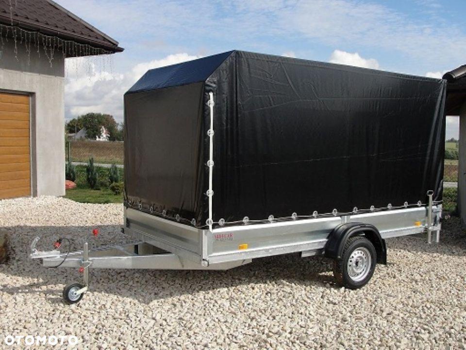 Przyczepa samochodowa z czarną plandeką, zaparkowana na żwirowym podłożu przed budynkiem z brązowymi drzwiami garażowymi.