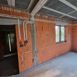 Wnętrze w stanie surowym z widoczną instalacją elektryczną w peszlach na ścianach z cegły dziurawki, z oknem i widokiem na zieleń za nim, oraz betonową wylewką na podłodze.