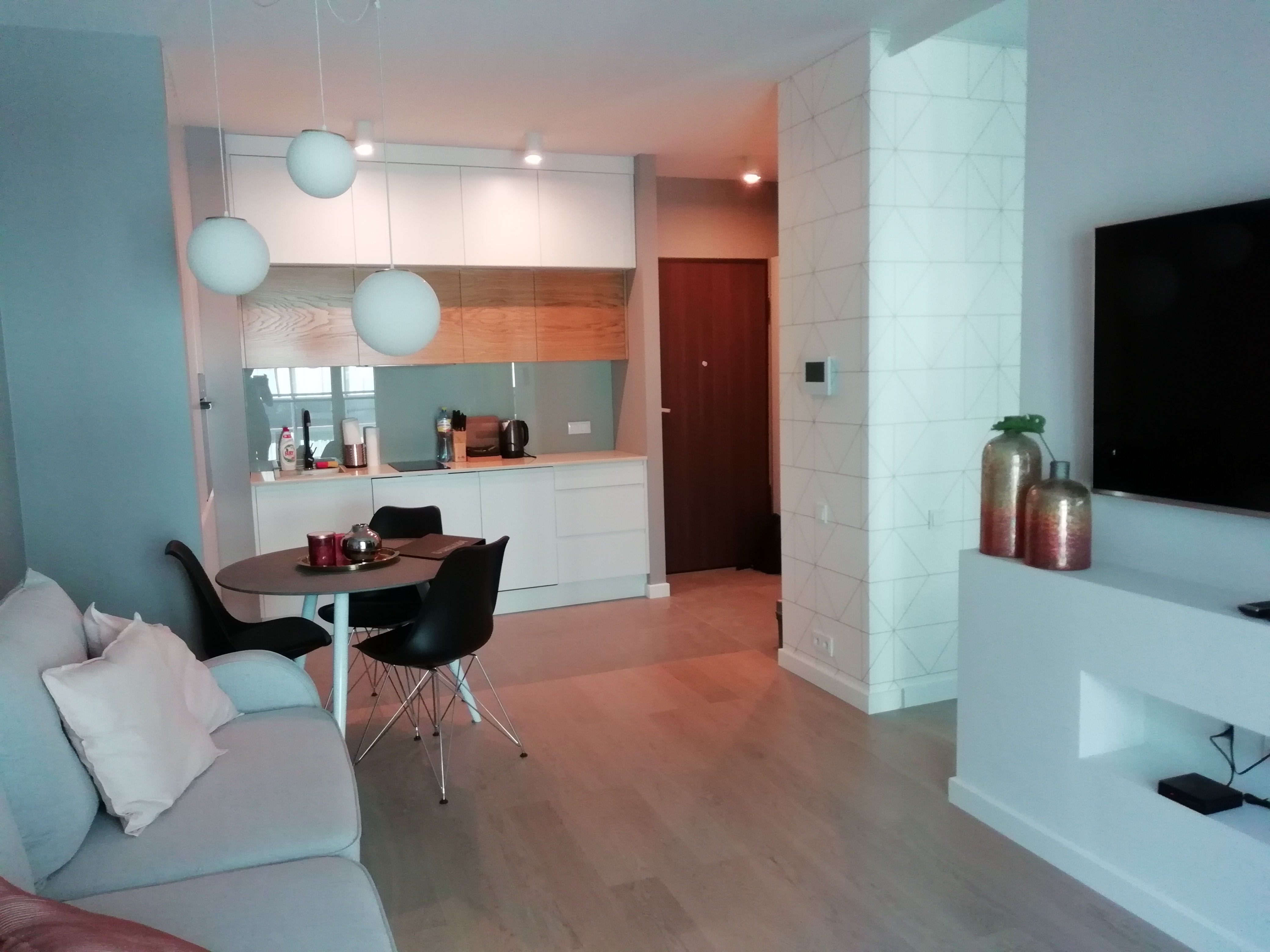 Nowoczesne wnętrze mieszkania z aneksem kuchennym, okrągłym stołem z czarnymi krzesłami i dekoracyjnymi lampami kulami. Ściana ozdobiona geometrycznym wzorem. Na komodzie dekoracyjne wazony.