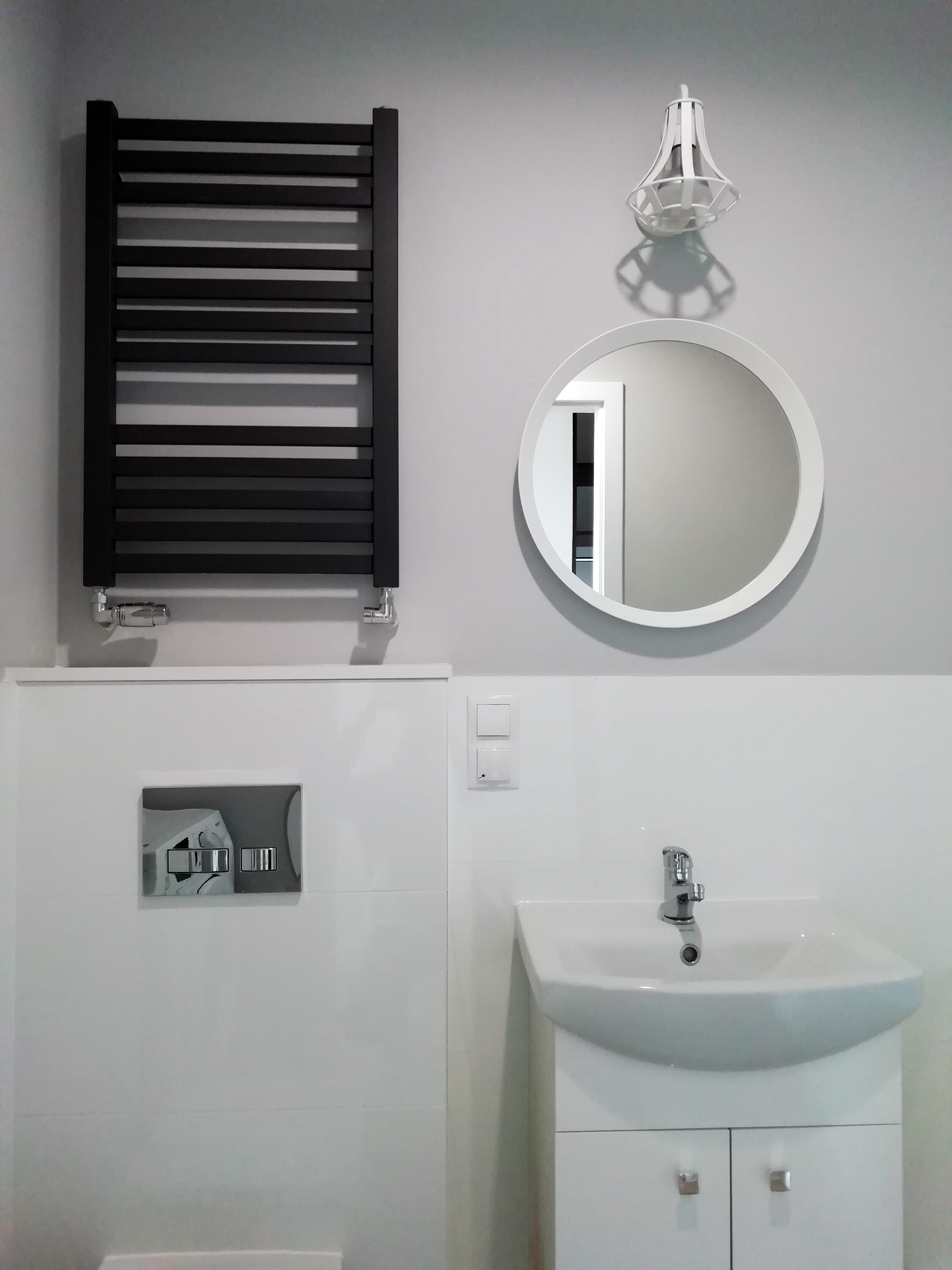 Nowoczesna łazienka w odcieniach szarości i bieli z czarnym grzejnikiem drabinkowym, okrągłym lustrem i minimalistyczną umywalką z szafką.
