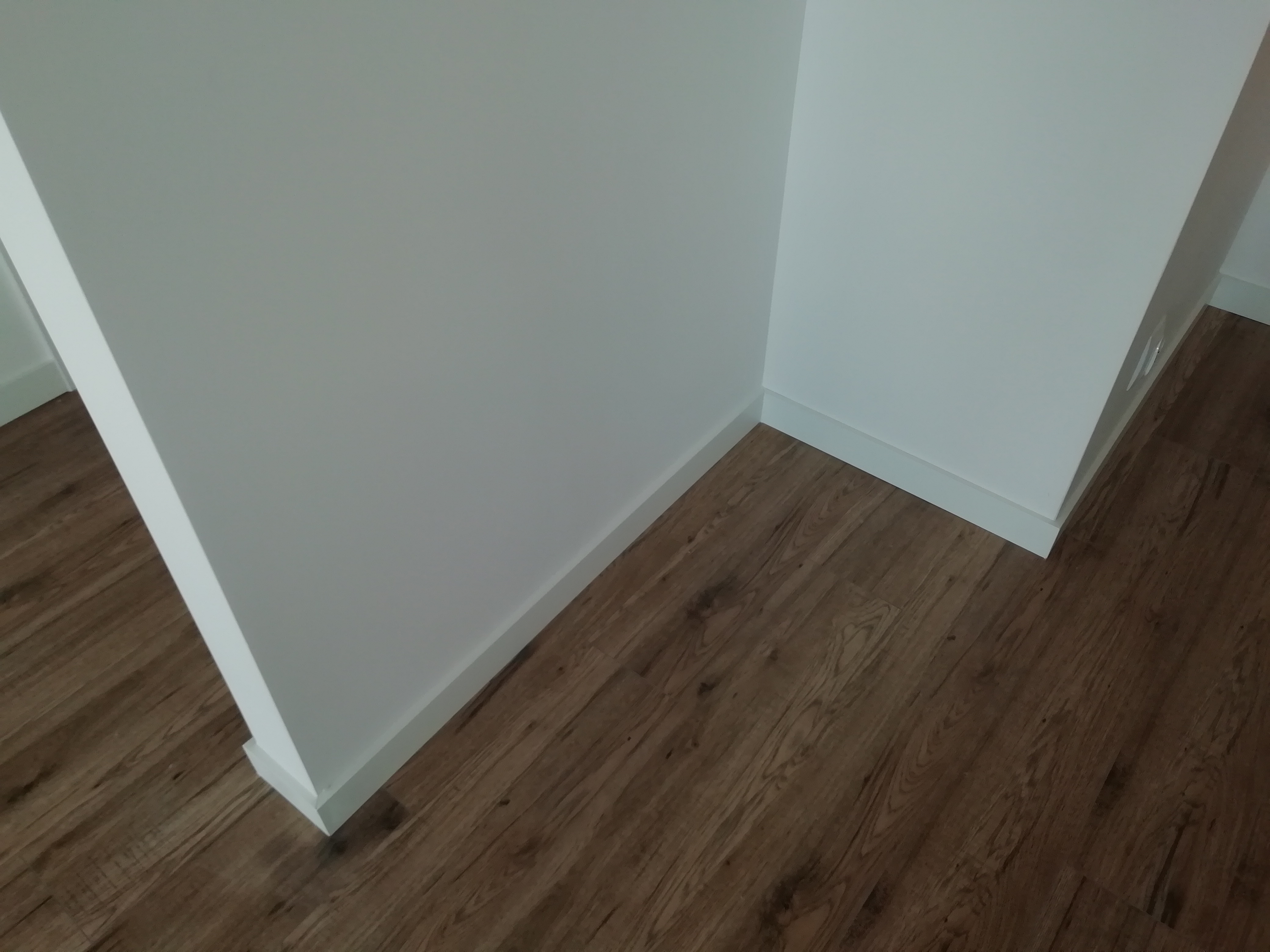 Wykończony narożnik pokoju z drewnianą podłogą i białymi ścianami z listwami przypodłogowymi, widoczne łączenie paneli podłogowych.