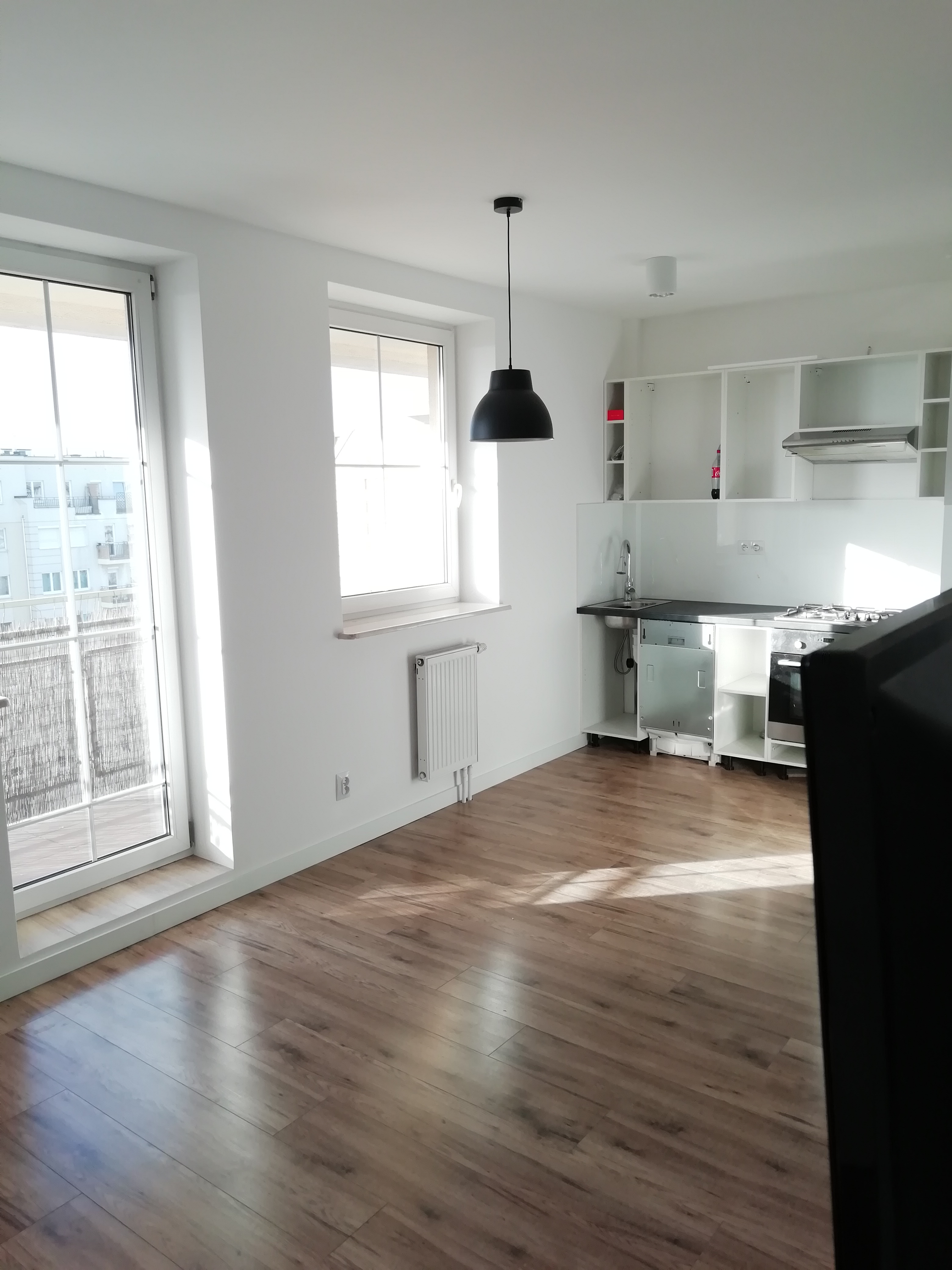 Jasne, przestronne wnętrze kuchni z drewnopodobną podłogą, białymi ścianami, oknem z widokiem na budynek mieszkalny i częściowo zamontowanymi białymi szafkami kuchennymi z czarnym blatem i sprzętem...