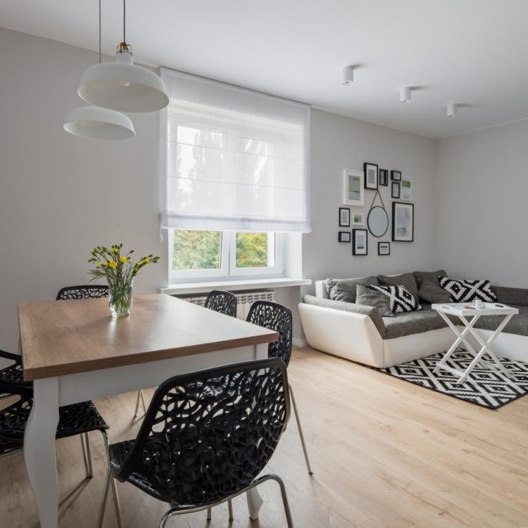 Jasne wnętrze pokoju dziennego z drewnianym stołem, czarnymi krzesłami o ażurowym wzorze, szarą sofą z geometrycznymi poduszkami i białą roletą rzymską na oknie.