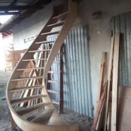 Niedokończone, spiralne schody z surowego drewna w warsztacie, z widocznymi narzędziami i materiałami budowlanymi w tle.