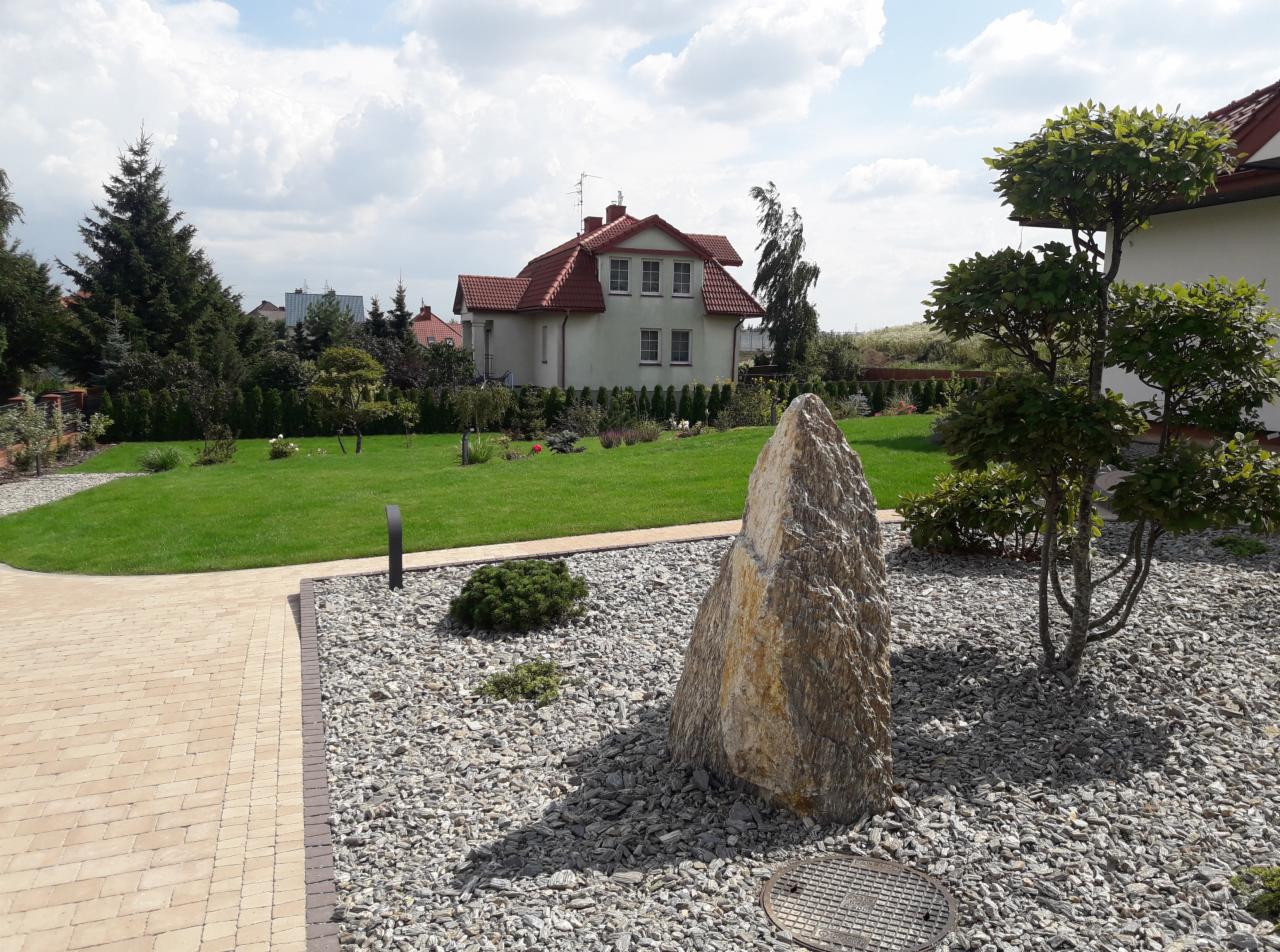 Uporządkowany ogród w Lublinie z trawnikiem, ozdobnymi krzewami i kamiennym monolitem na tle domu jednorodzinnego.