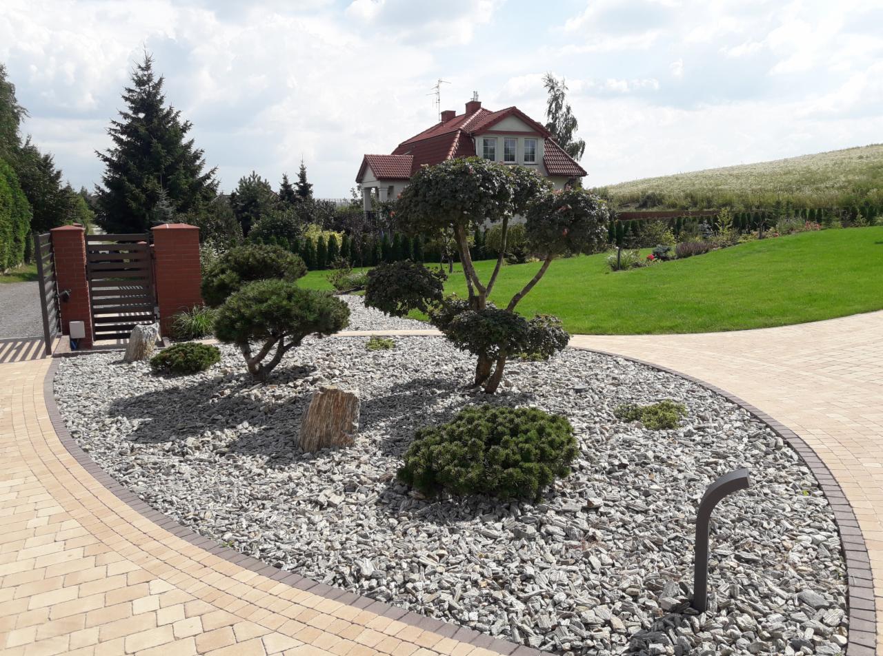 Ogród z ozdobnymi drzewami formowanymi, wysypany grysem, z kostką brukową i widokiem na dom w tle.