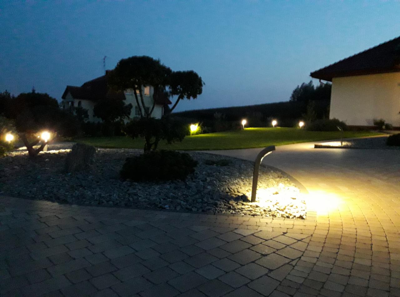 Oświetlenie ogrodu po zmierzchu: lampy punktowe podkreślają ścieżkę z kostki brukowej, trawnik i ozdobne krzewy.