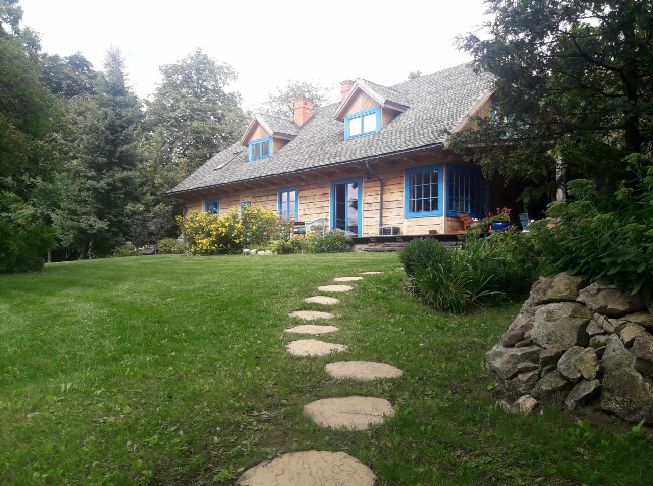 Drewniany dom z niebieskimi oknami i kamienną ścieżką przez trawnik, otoczony zielenią i drzewami.