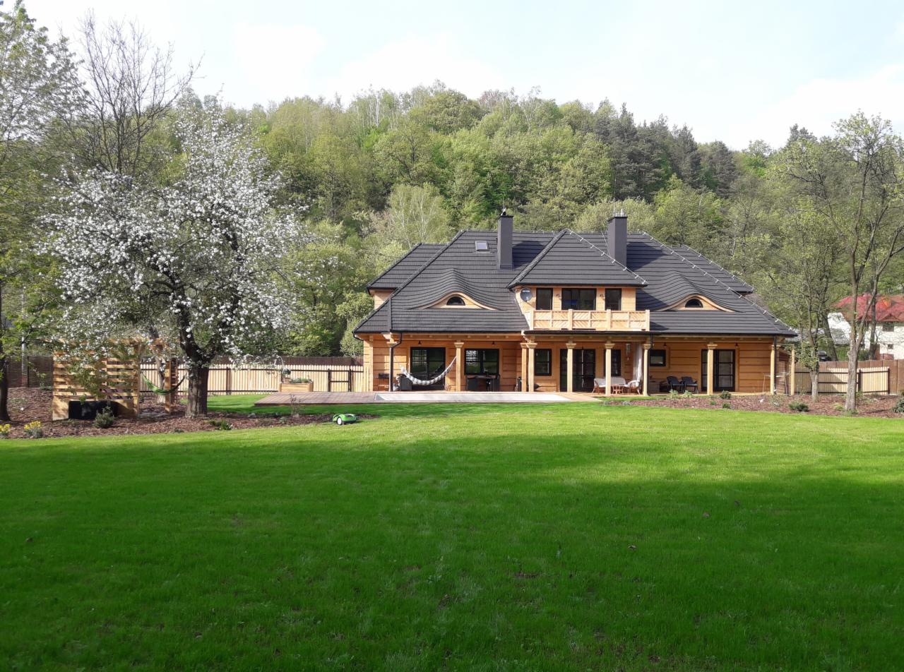 Duży, drewniany dom z czarnym dachem, kolumnami i tarasem, otoczony zielonym trawnikiem i kwitnącym drzewem owocowym.