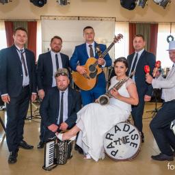 Zespół Paradise z Leszna Wielkopolska - Zespół muzyczny w strojach formalnych, w tym panna młoda z saksofonem, pozuje z gitarą, akordeonem i marakasami przed sprzętem nagłośnieniowym na tle sali z dekoracjami.