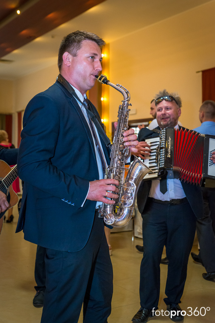 Saksofonista w granatowym garniturze gra na srebrnym saksofonie tenorowym, w tle akordeonista i inni goście na uroczystości.
