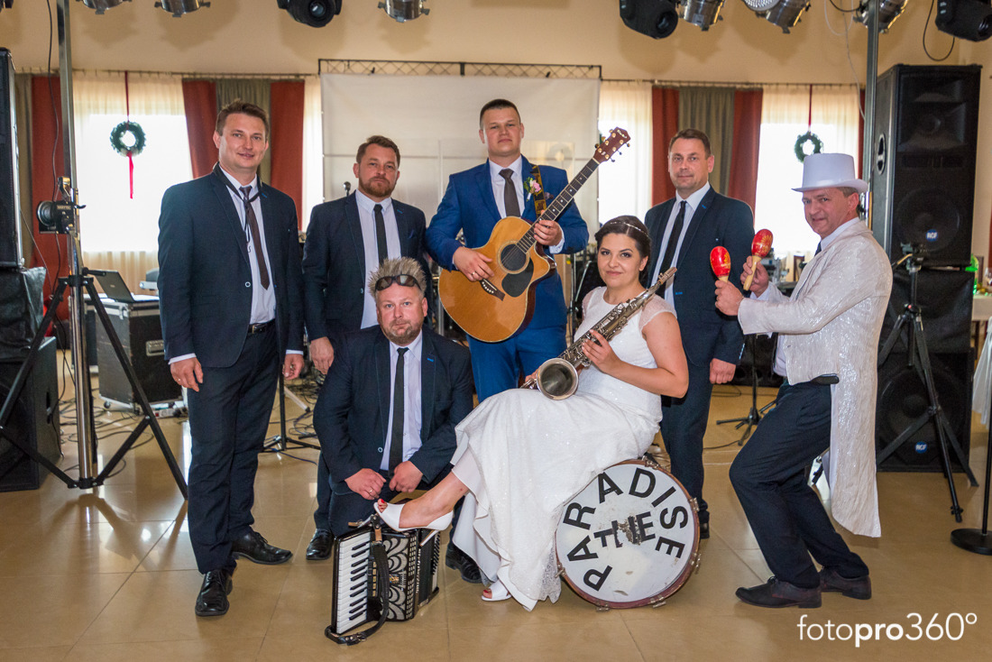 Zespół muzyczny w strojach formalnych, w tym panna młoda z saksofonem, pozuje z gitarą, akordeonem i marakasami przed sprzętem nagłośnieniowym na tle sali z dekoracjami.