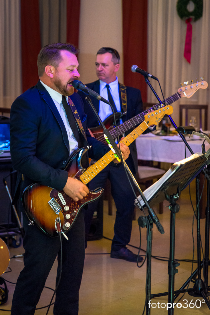 Wokalista w ciemnym garniturze śpiewa do mikrofonu, grając na gitarze elektrycznej typu Stratocaster z brązowo-żółtym wykończeniem, w tle drugi gitarzysta basowy i dekoracja z zielenią.