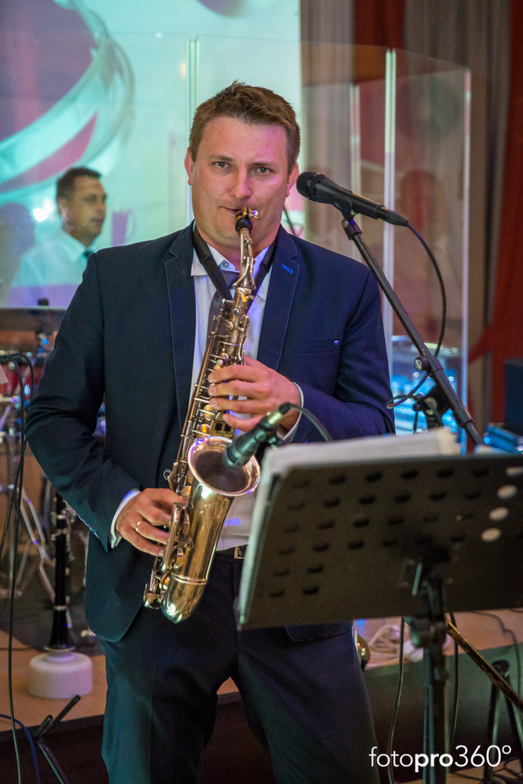 Saksofonista w ciemnym garniturze gra na saksofonie podczas występu na scenie, widoczny stojak na nuty i mikrofon.