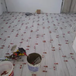 Układanie jasnych paneli podłogowych z użyciem czerwonych klipsów poziomujących; widoczne narzędzia: poziomica, wiadro z zaprawą i wiadro z klipsami.