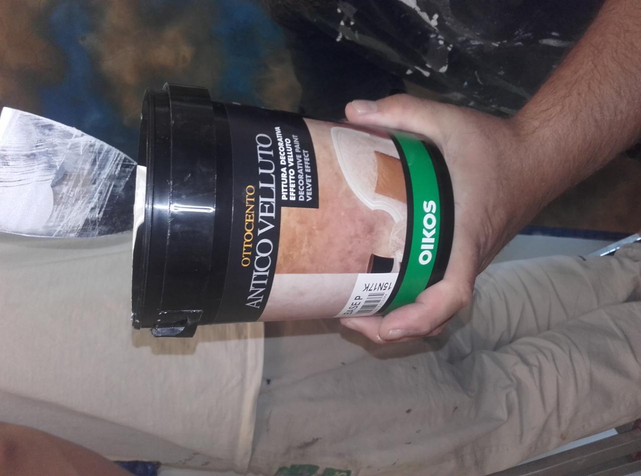 Otwarta puszka farby dekoracyjnej Ottocento Antico Velluto z logo Oikos, szpachelka z resztkami białej farby, widoczne spodnie robocze malarza