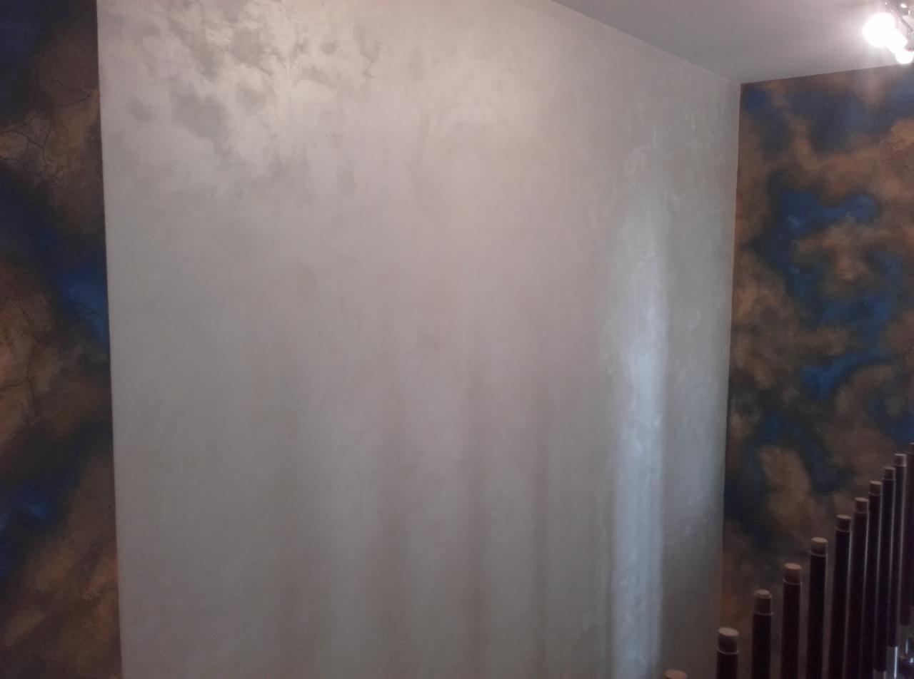 Ściana wykończona dekoracyjnym tynkiem w odcieniach szarości z subtelnym połyskiem, sąsiadująca z fragmentem ściany pokrytej wzorem imitującym marmur w kolorach brązu i błękitu, widoczne elementy...