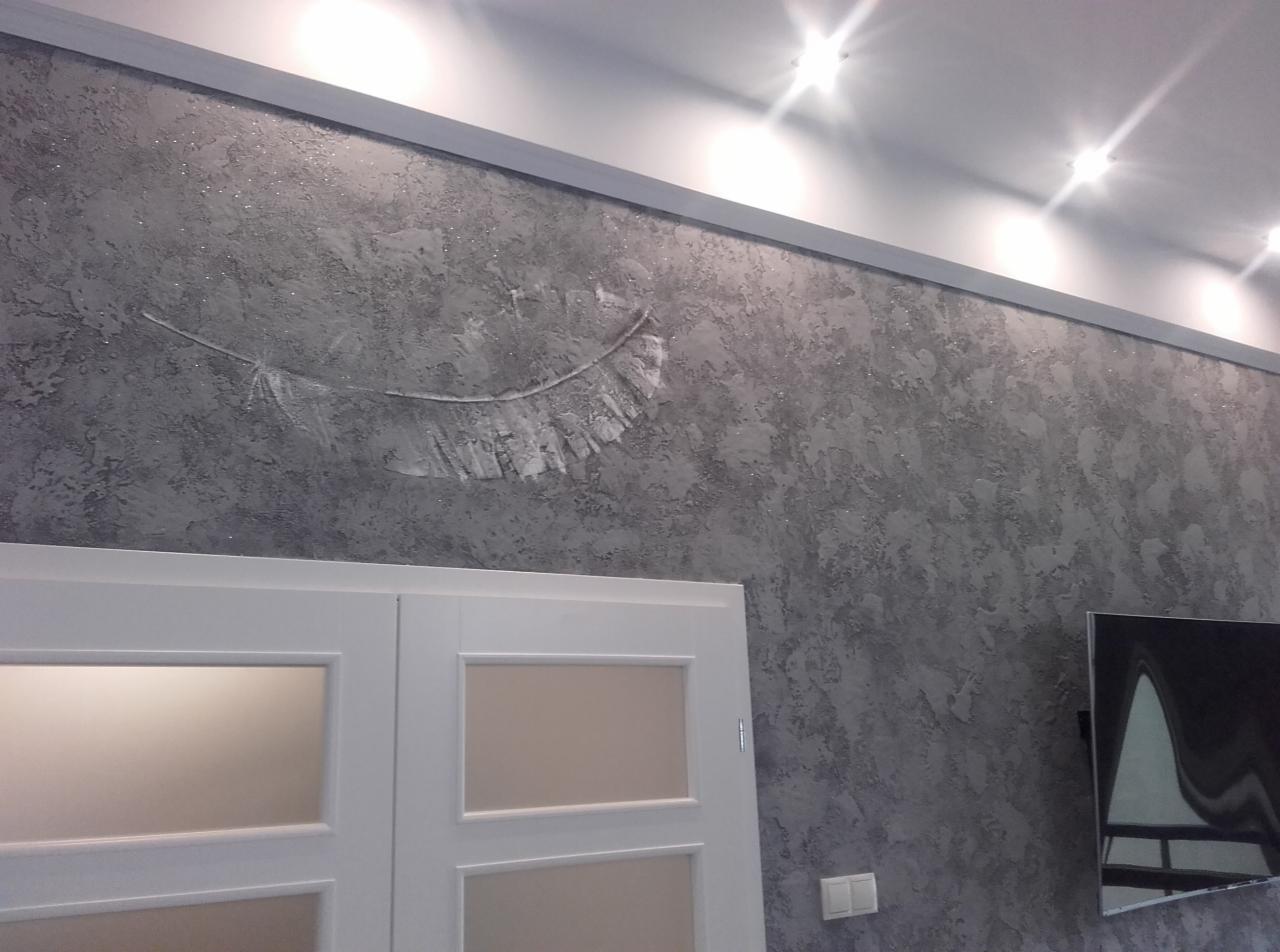 Dekoracyjna ściana w odcieniach szarości z efektem struktury i srebrnym motywem pióra, widoczny fragment białych drzwi i telewizora.