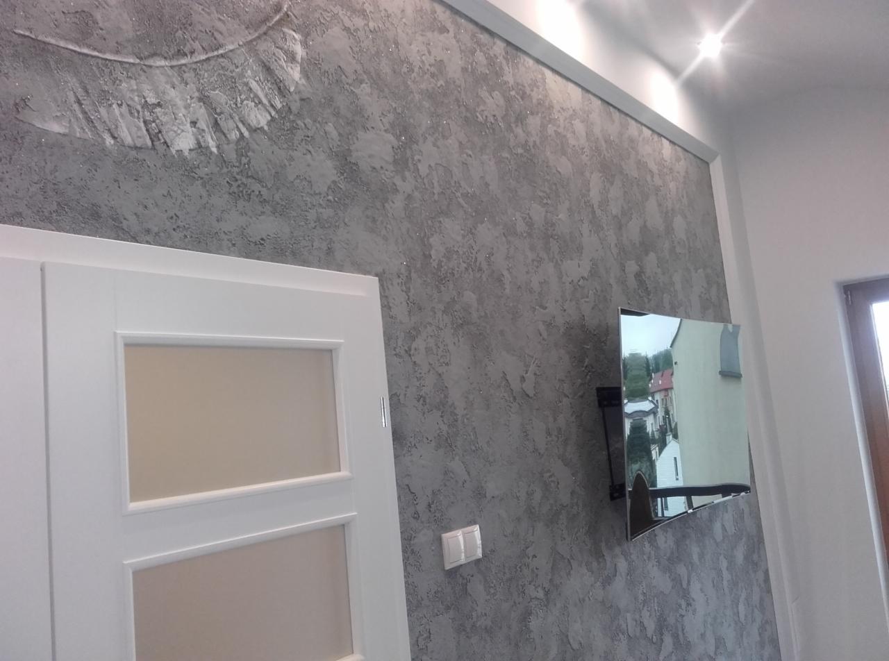 Ściana z dekoracyjnym tynkiem w odcieniach szarości z motywem pióra, widoczna w mieszkaniu w Świdniku, obok białe drzwi z przeszkleniami i zawieszony telewizor.