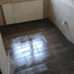 Świeżo ułożona, mokra, ciemnobrązowa podłoga z płytek imitujących drewno w narożnej łazience, widoczny grzejnik, okno i fragment ściany z kranem.