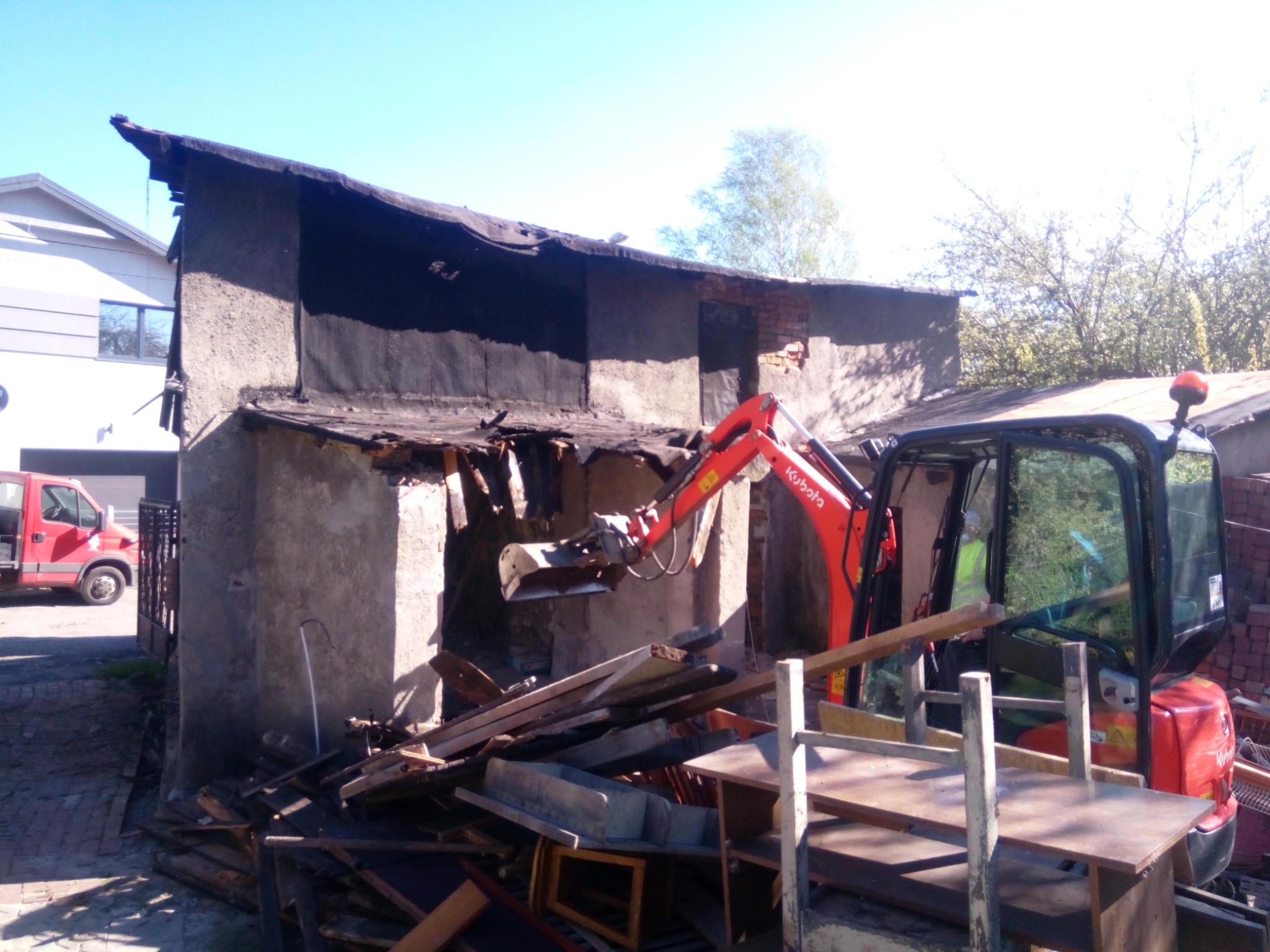 Częściowo zburzony budynek gospodarczy z czerwonym mini-koparką Kubota w trakcie prac rozbiórkowych, widoczne gruz i stare meble, w tle budynek mieszkalny i czerwony samochód dostawczy.
