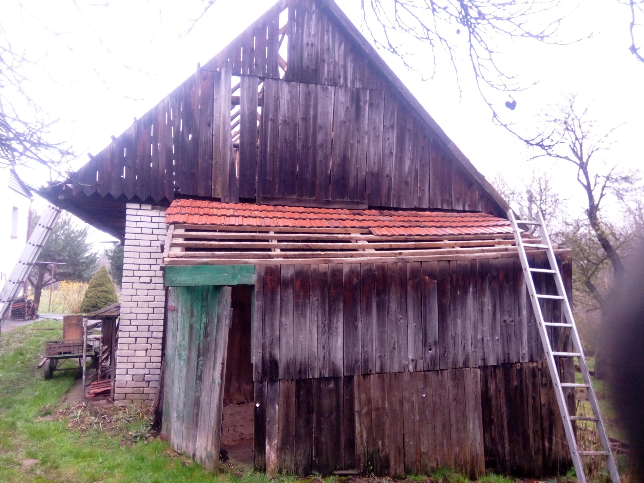 Drewniana stodoła z częściowo zdjętym dachem, odsłonięta konstrukcja więźby dachowej, drabina oparta o ścianę, widoczne ślady demontażu.