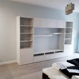 Biała, nowoczesna meblościanka z półkami i szafkami, zamontowana na tle jasnej ściany w pokoju z drewnianą podłogą. Na pierwszym planie widoczne elementy wyposażenia wnętrza.