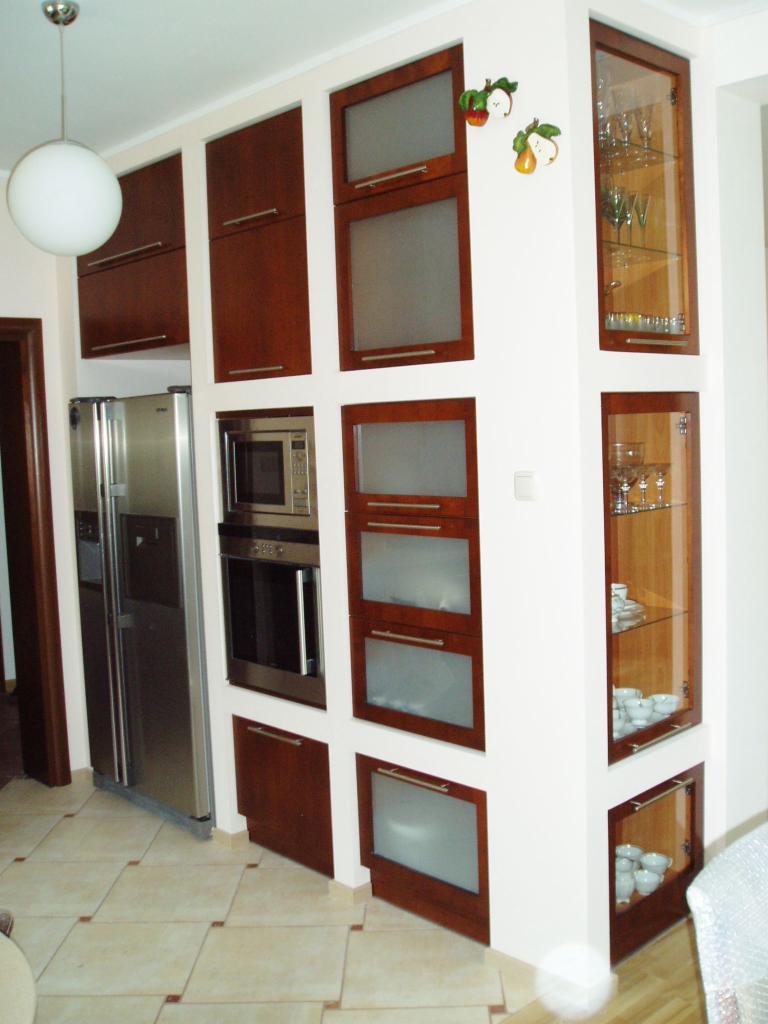 Kuchnia z zabudową meblową w odcieniach brązu i beżu, wyposażona w sprzęt AGD: lodówkę, piekarnik i mikrofalówkę. Na ścianie dekoracja w kształcie jabłek.