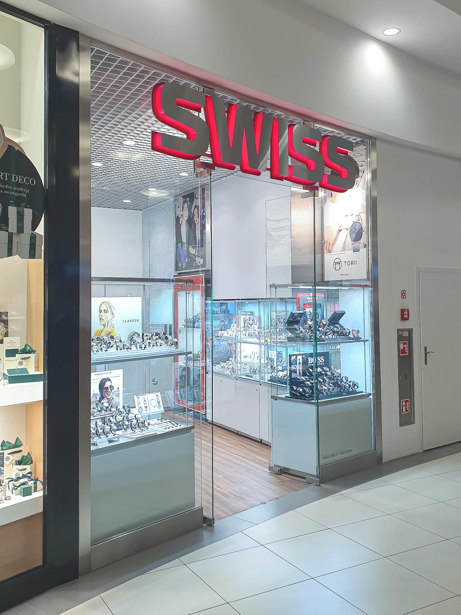 Wejście do butiku z zegarkami Swiss z podświetlanym na czerwono napisem, przeszklone witryny eksponują zegarki różnych marek, w tle widoczna ściana z ekspozycją zegarków.