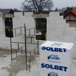 Wznoszenie ścian domu z bloczków Solbet, widoczne rusztowanie, drewniane szalunki nad otworami okiennymi, w tle zabudowa jednorodzinna.