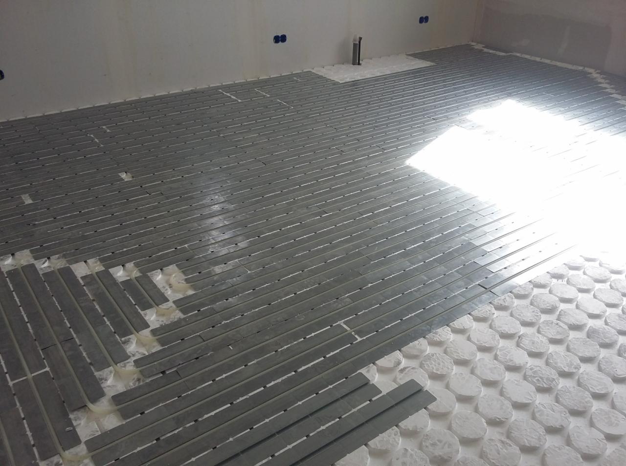 Montaż szarej, podłużnej mozaiki na podłodze z ogrzewaniem podłogowym; widoczne białe elementy systemu ogrzewania podłogowego i częściowo ułożona mozaika.