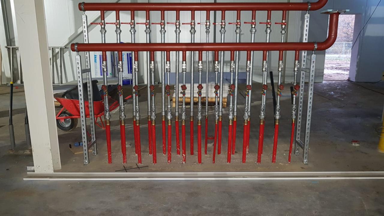 Instalacja centralnego ogrzewania: czerwone rury pionowe i poziome z zaworami, zamontowane na metalowej konstrukcji w surowym wnętrzu budynku.