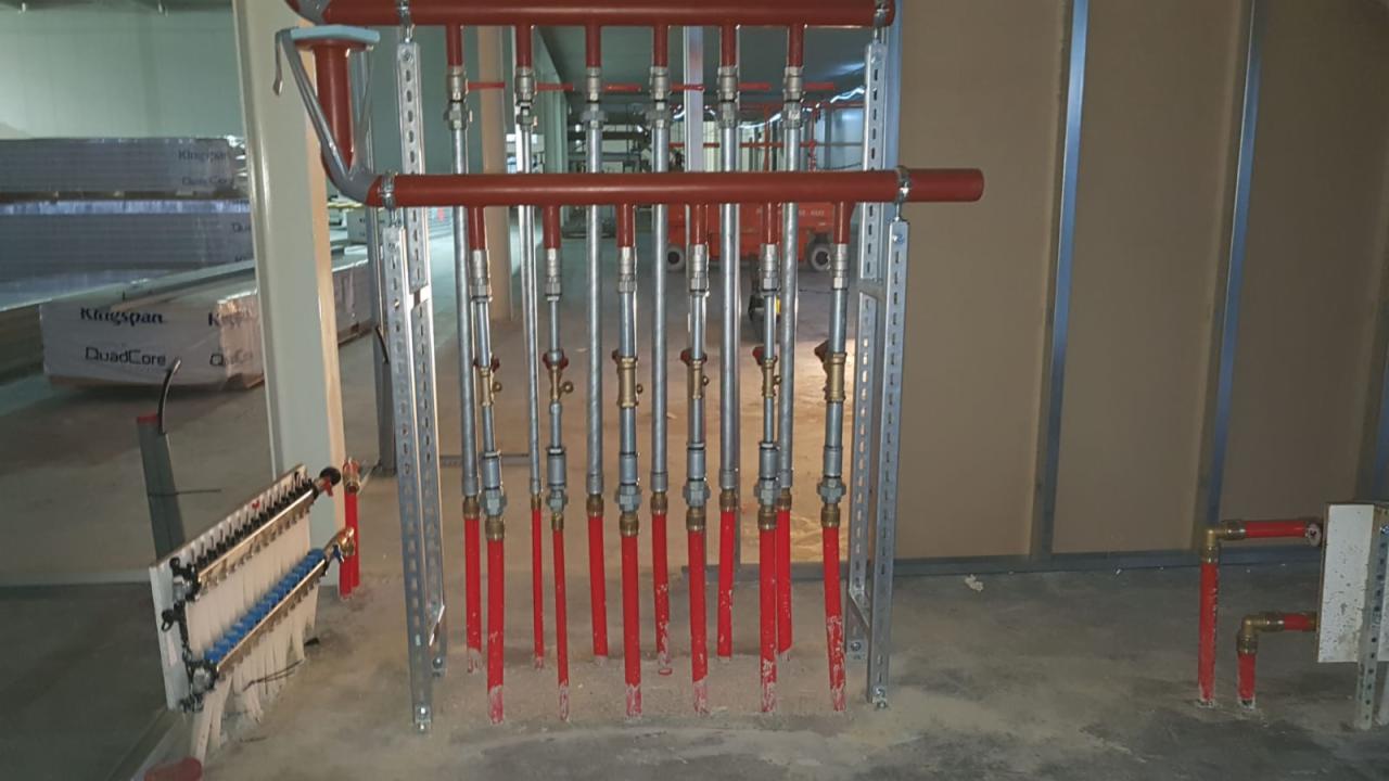 Instalacja centralnego ogrzewania z czerwonymi rurami, srebrnymi zaworami i metalowymi wspornikami, widoczna w surowym stanie budynku, z paczkami materiałów budowlanych w tle.