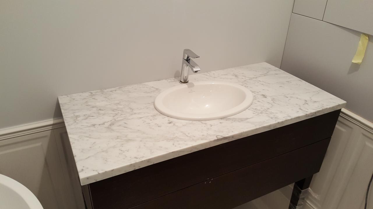 Umywalka nablatowa wpuszczona w blat z białego marmuru z szarymi żyłami, zamontowana na ciemnobrązowej szafce łazienkowej z widoczną baterią.