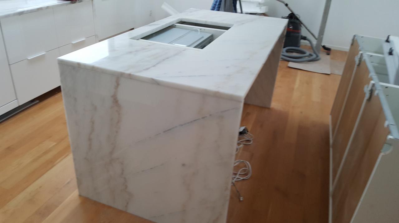 Biały blat kuchenny z marmurowym wzorem, z wycięciem na płytę grzewczą, w trakcie montażu, widoczne szafki kuchenne i sprzęt do sprzątania.
