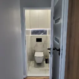 Nowoczesna toaleta w odcieniach szarości i bieli, widoczna z korytarza przez uchylone białe drzwi z czarnymi klamkami, z podświetlanymi szafkami nad sedesem i marmurowymi płytkami na ścianach.