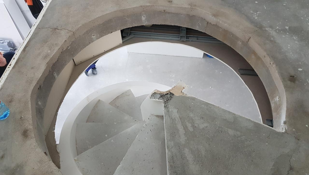 Widok z góry na betonowe schody spiralne w trakcie budowy, z widoczną surową konstrukcją i elementami szalunku, oraz fragmentem pracownika w tle.