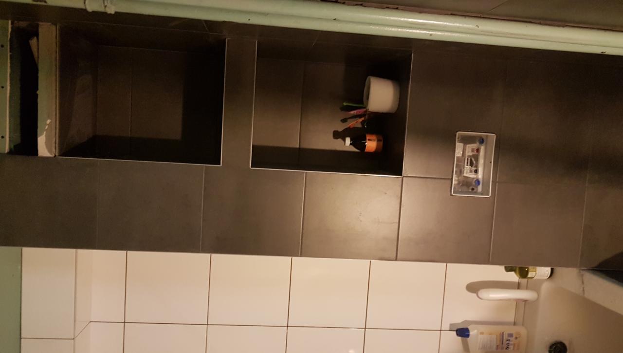 Ściana łazienki z szarymi płytkami i wnękami, w jednej z nich papier toaletowy i butelka, poniżej białe płytki nad wanną.