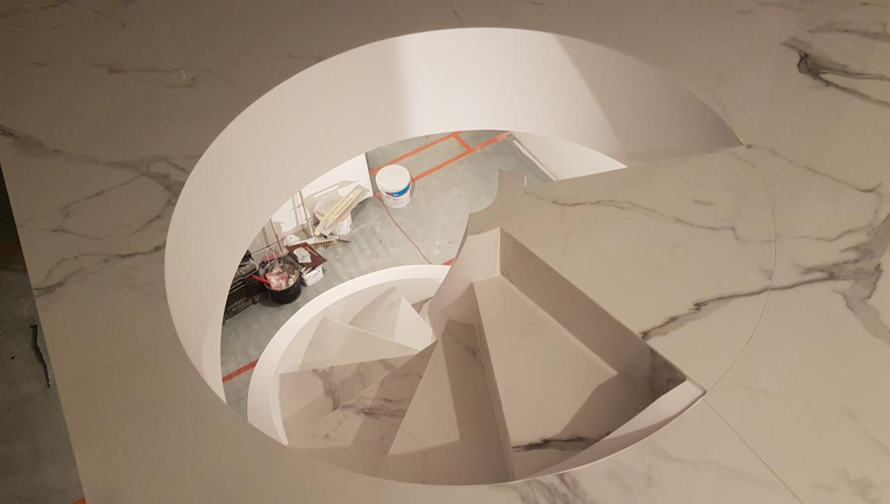 Widok z góry na spiralne schody wyłożone płytkami imitującymi marmur, w trakcie montażu, widoczne narzędzia i wiadro z farbą na podłodze poniżej.