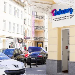 Fasada budynku z szyldem 'Shakeaway' w słoneczny dzień, widok z ulicy z zaparkowanymi samochodami, w tle inne budynki i szyldy reklamowe.