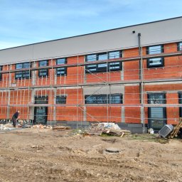 Alu-mar klaudia paluch - Budynek w trakcie ocieplania, widoczne rusztowania, okna i cegły. Pracownik w dolnej części kadru. Na ziemi gruz i materiały budowlane.