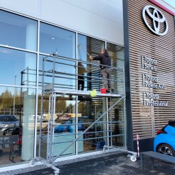 Alu-mar klaudia paluch - Pracownik myje okna na rusztowaniu przed salonem Toyoty. Widoczne logo i napisy informacyjne. Jasne, duże okna i czyste otoczenie.