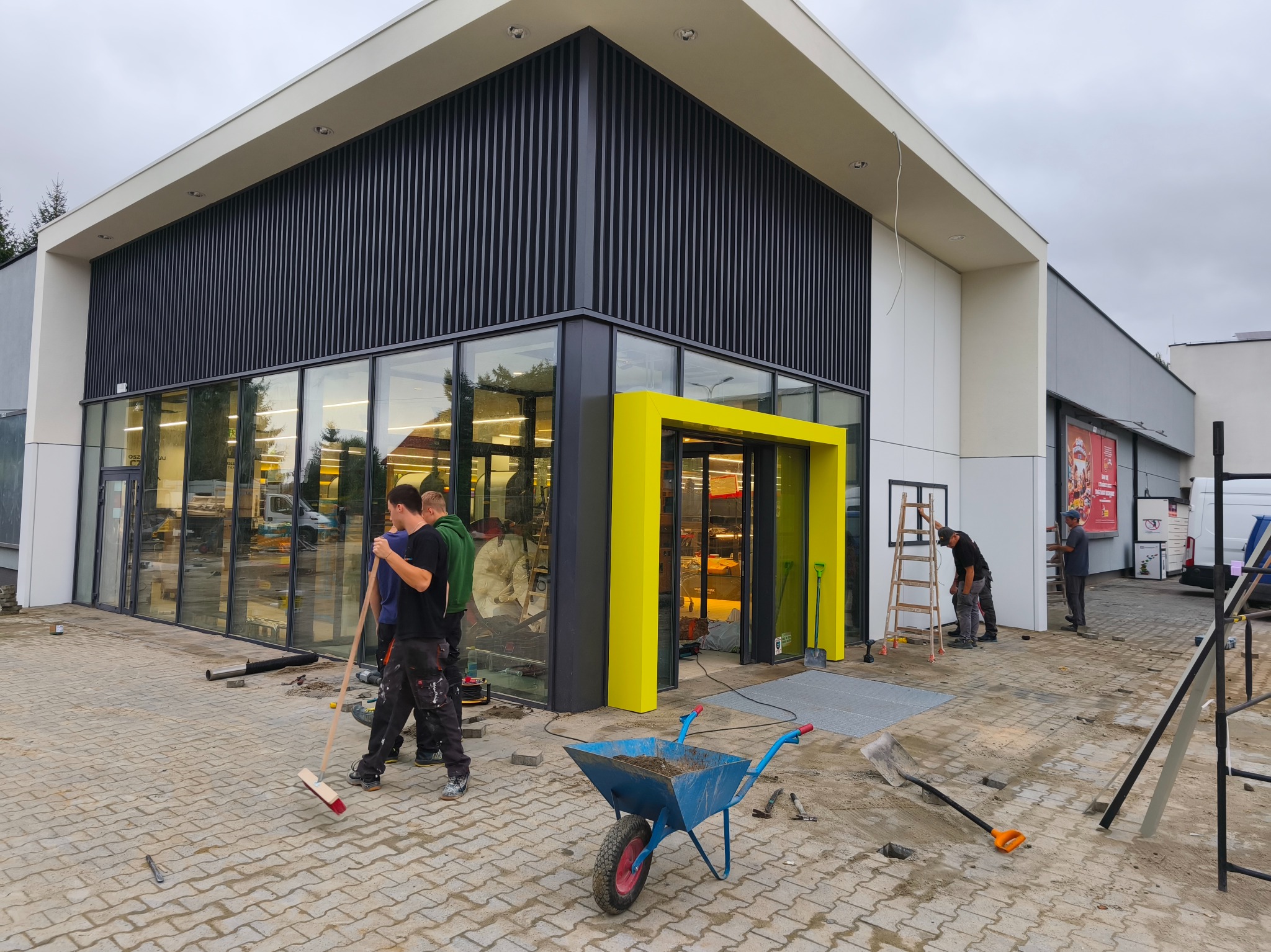 Montaż witryny sklepowej z żółtym wejściem. Pracownicy sprzątają teren budowy, widoczny wózek z ziemią i narzędzia. Nowoczesna architektura budynku.