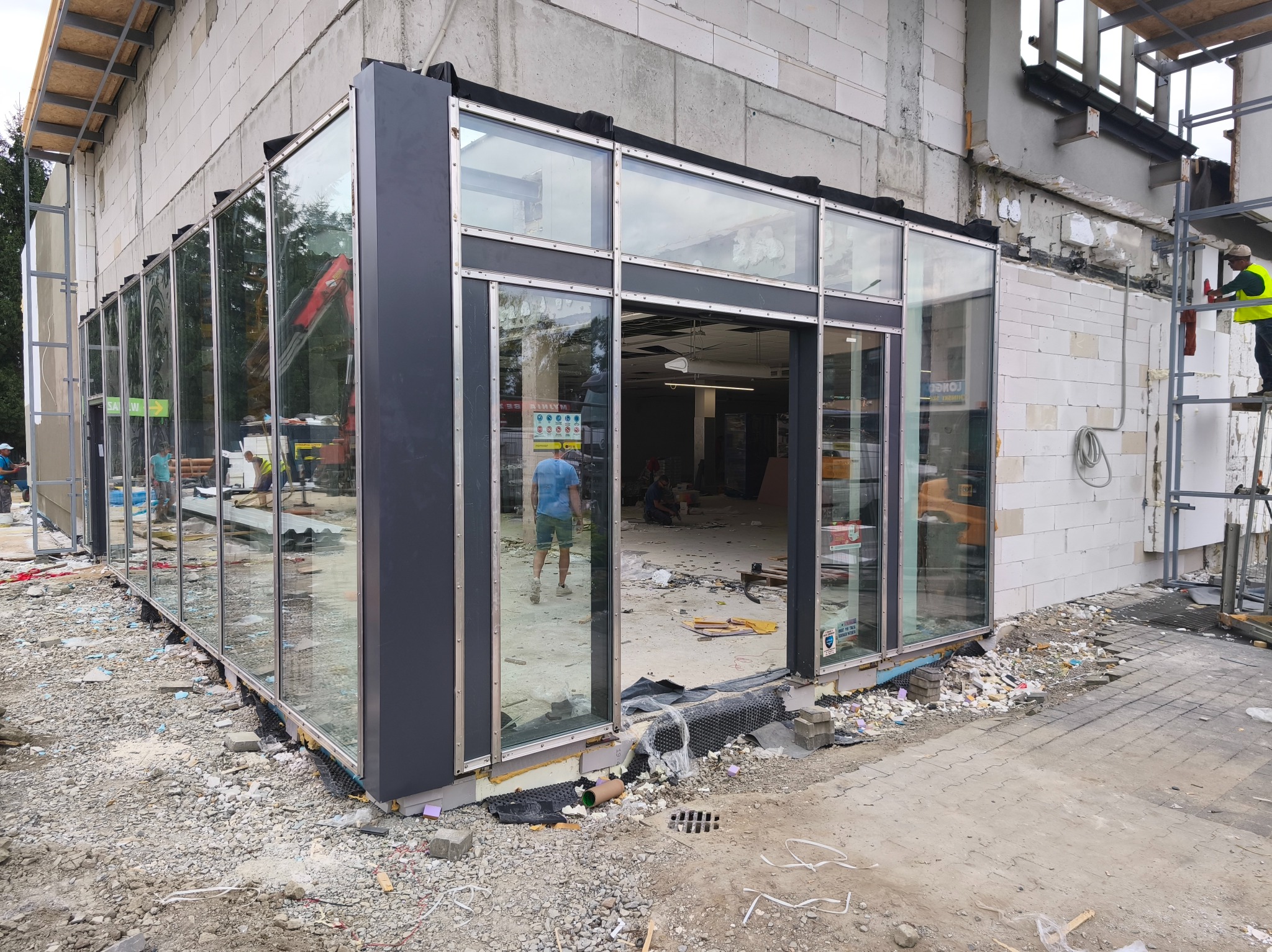 Montaż szklanej fasady budynku w trakcie prac wykończeniowych. Widoczne rusztowania i materiały budowlane. Częściowo wykończone wnętrze.