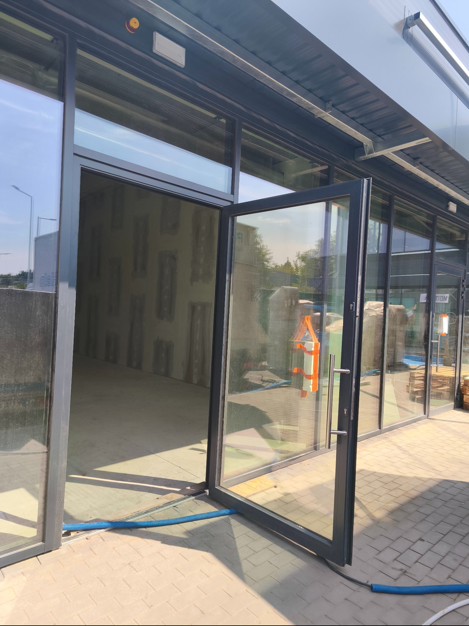 Otwarta, przeszklona, nowoczesna aluminiowa witryna drzwiowa z widocznym wnętrzem budynku w surowym stanie. Na zewnątrz kostka brukowa i niebieski wąż.